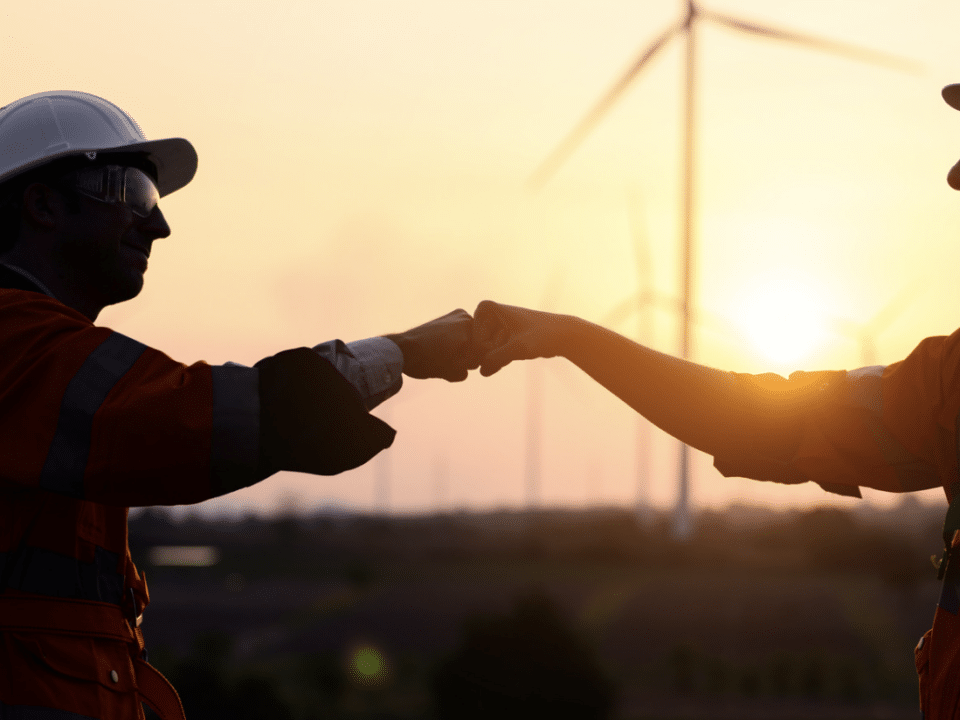 two silhouetted wind turbine technicians give fist bumps in front of wind farm